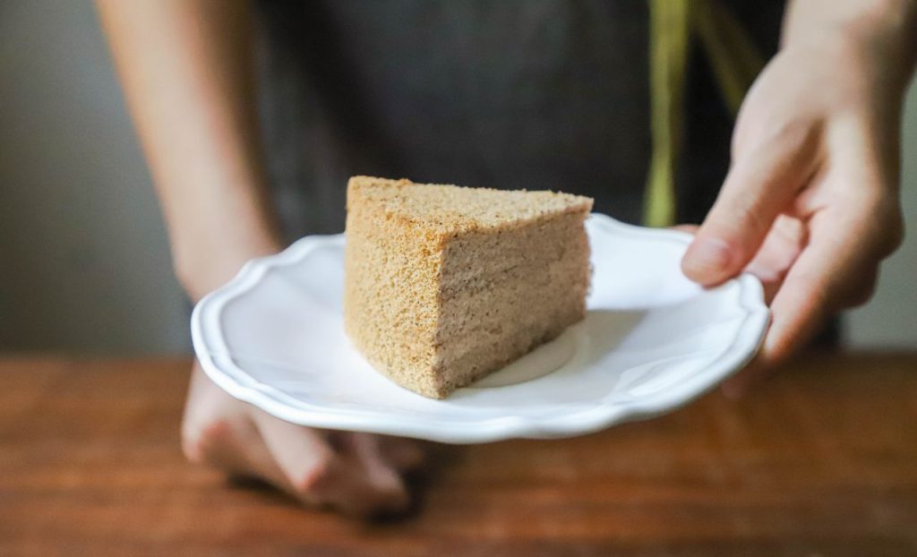 Hand holding a piece of homemade cake on a white plate, offering it as a treat.