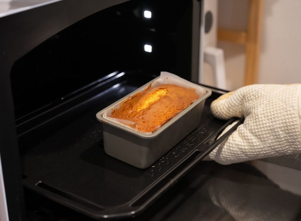 A loaf cake being taken out of an oven using a white oven mitt, capturing the warmth of home baking.
