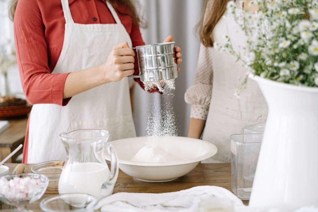 Two women in aprons sifting flour with milk in a cozy kitchen setting.