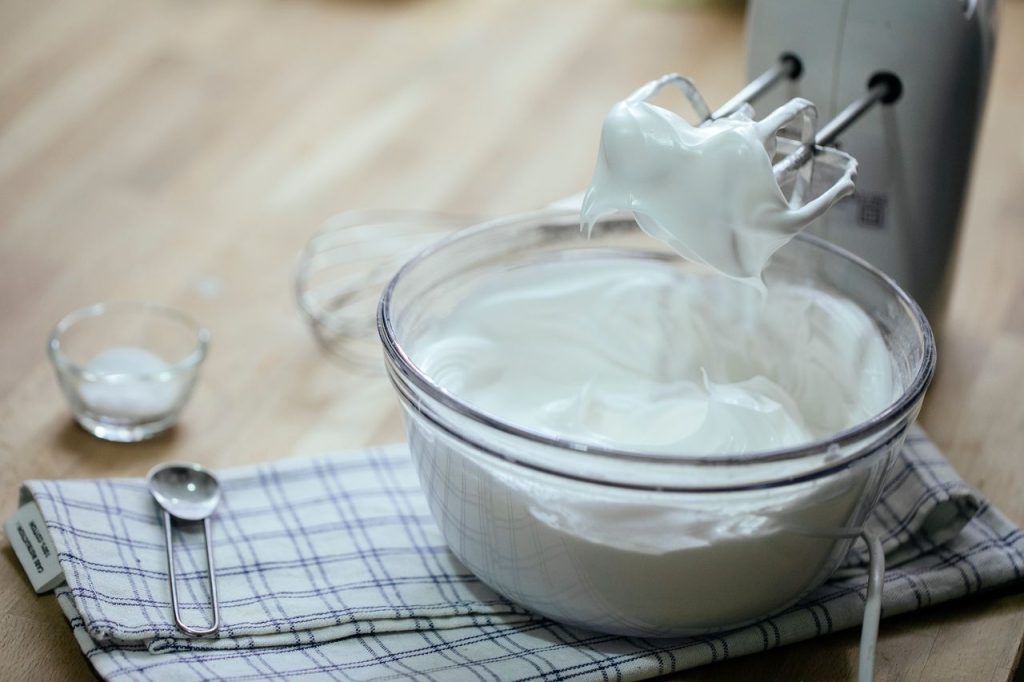 Arrangement of tasty fresh whipped cream in glass bowl near mixer spoon on towel on kitchen table